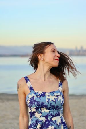 Chantelle's profile photo which is a person flipping their should length dark hair over their shoulder. They are wearing a blue and white tropical botanical patterned dress. They are at one of their favourite places which is the beach with the so-called city of vancouver off in the distance